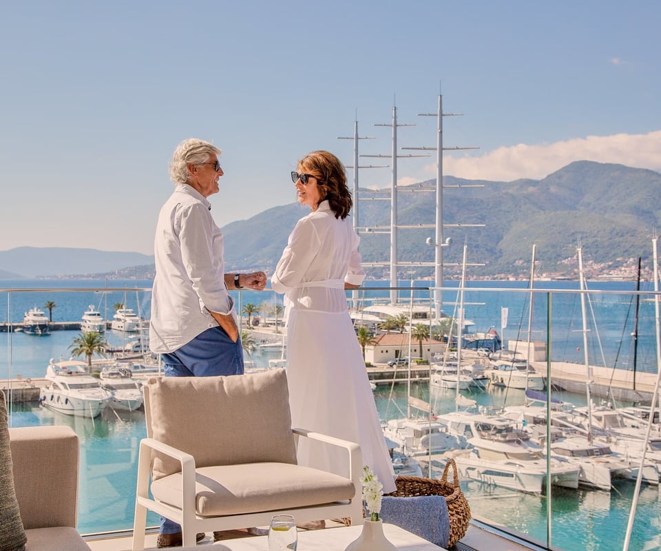 A couple standing on a balcony with a panoramic view of a marina filled with yachts and sailboats, with mountains in the background under a clear blue sky.