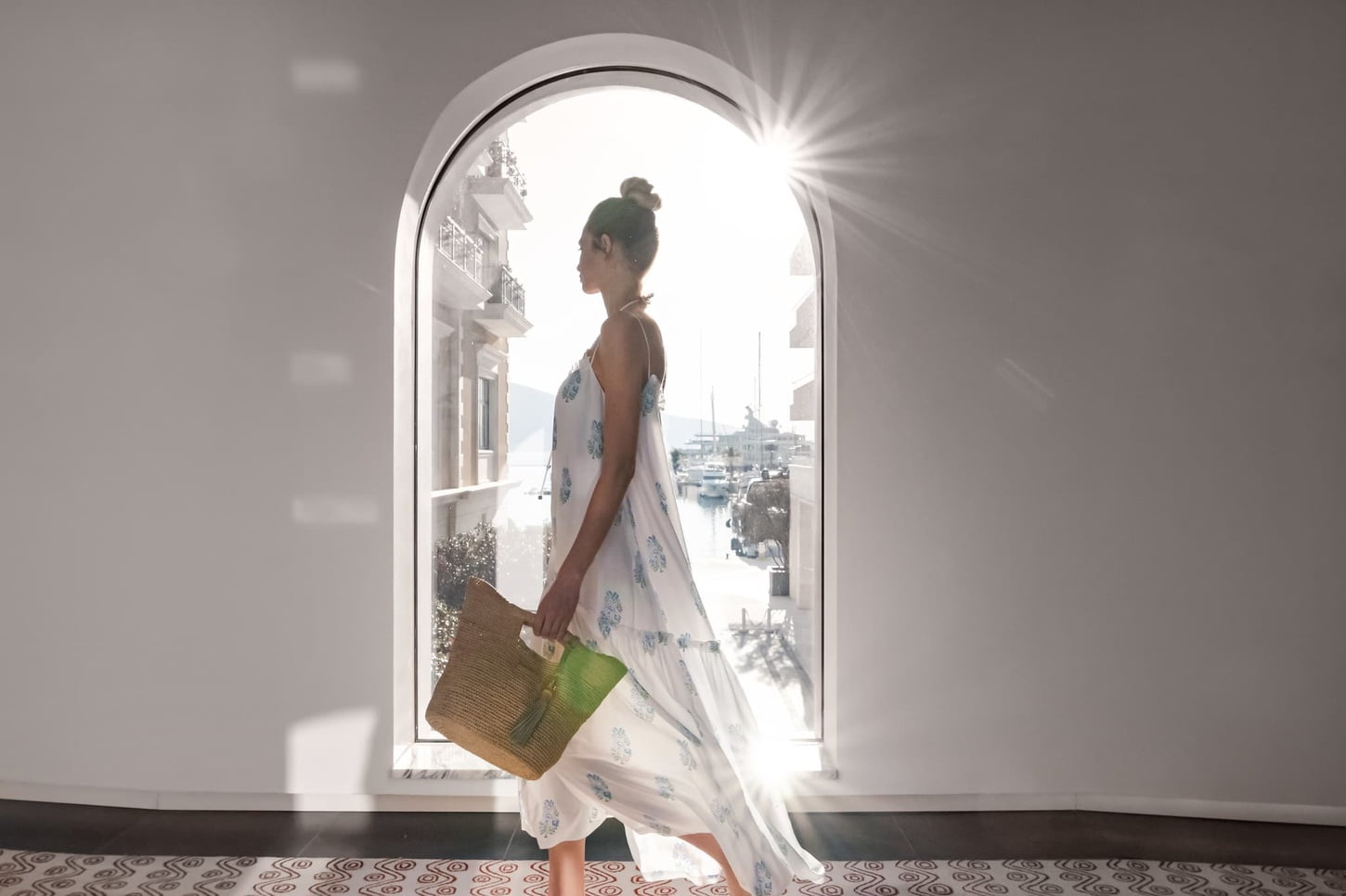 A woman in a flowing white dress with blue patterns stands in a sunlit arched doorway holding a straw tote bag.