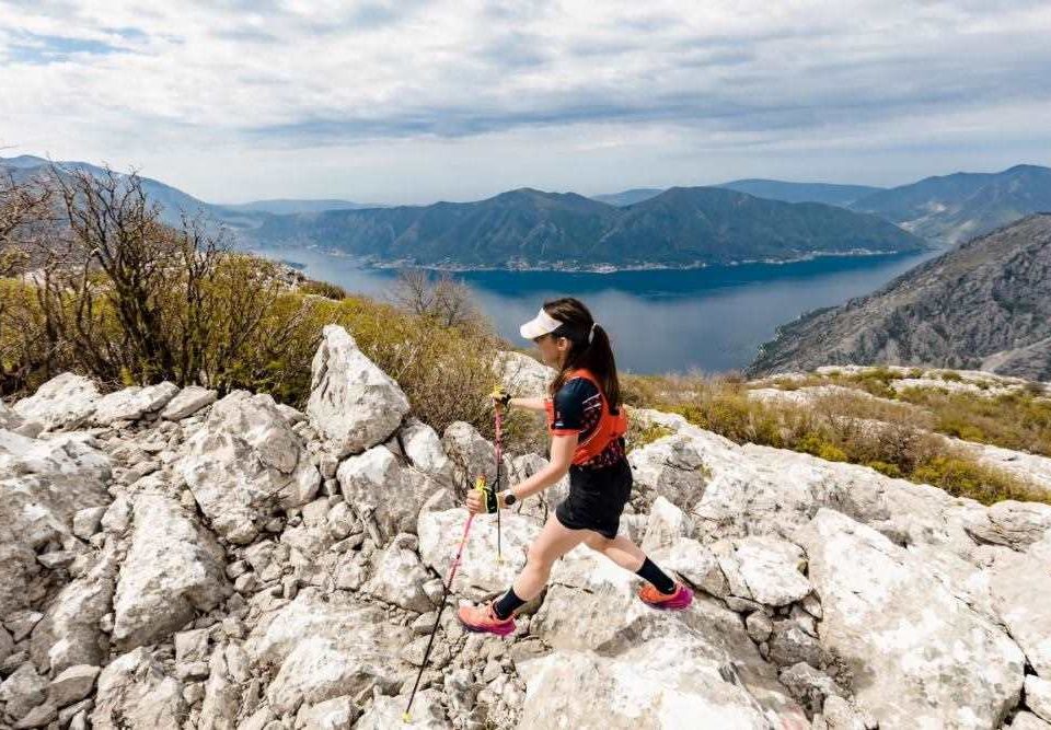 A female trail runner wearing a visor, sunglasses, and athletic gear is navigating a rocky mountain path with trekking poles. She is surrounded by rugged terrain with large white stones, sparse vegetation, and breathtaking views of the Boka Bay in Montenegro. The deep blue waters of the bay contrast against the towering green mountains in the background under a partly cloudy sky.