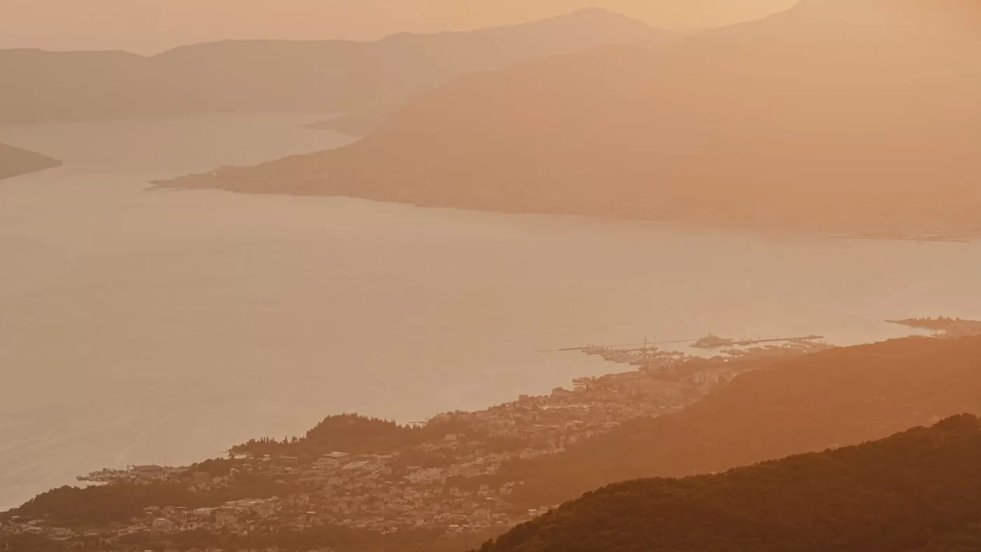 Serene aerial view of Boka Bay at sunset, with warm golden light casting a soft glow over the tranquil waters and surrounding mountains, creating a peaceful, ethereal landscape.