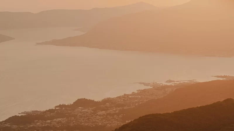 Serene aerial view of Boka Bay at sunset, with warm golden light casting a soft glow over the tranquil waters and surrounding mountains, creating a peaceful, ethereal landscape.