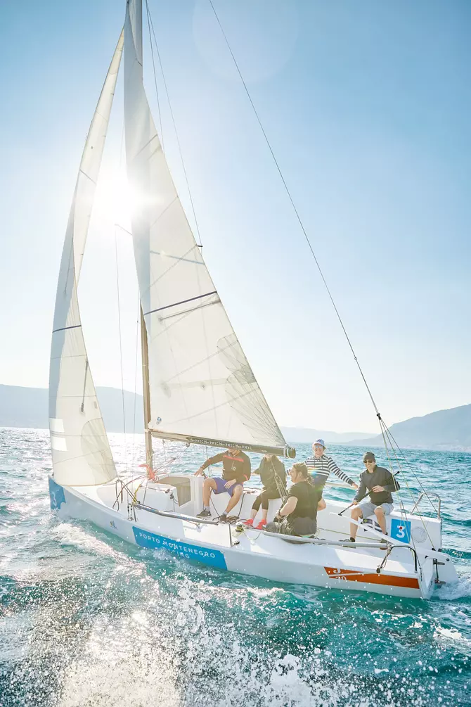 Five people navigating a sailboat under bright sunlight, with the sun casting a glow through the sails and the vibrant blue sea creating a stunning backdrop