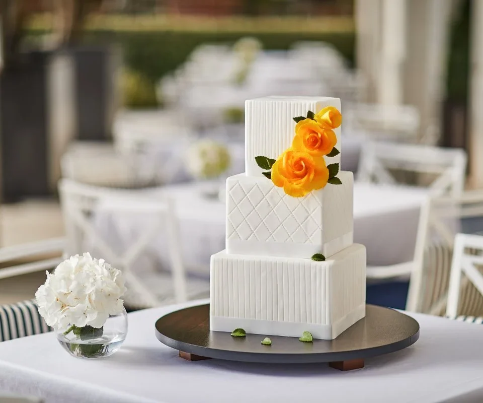 Elegant three-tiered square cake with white fondant textures, adorned with yellow roses. It sits on a dark serving board, with a vase of white flowers nearby.
