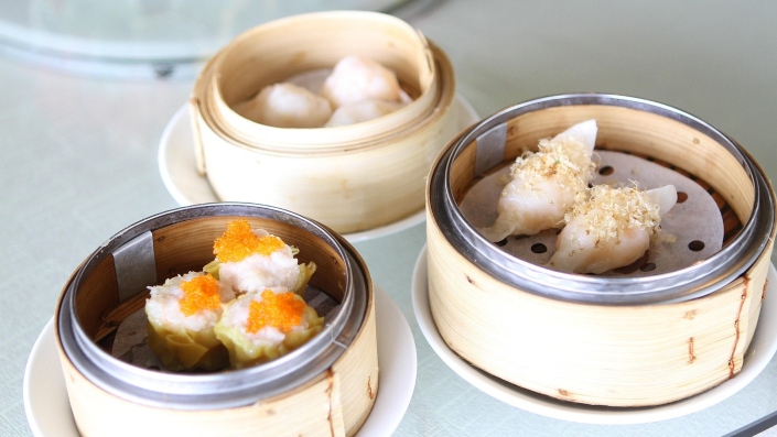 Three bamboo steamers filled with different types of dim sum: shrimp dumplings, siu mai topped with orange roe, and translucent dumplings garnished with dried shrimp.