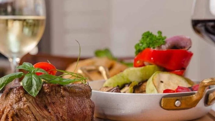 A succulent steak with mixed grilled vegetables served on a wooden board, paired with a glass of white wine - representing the modern Adriatic brasserie One at Porto Montenegro.