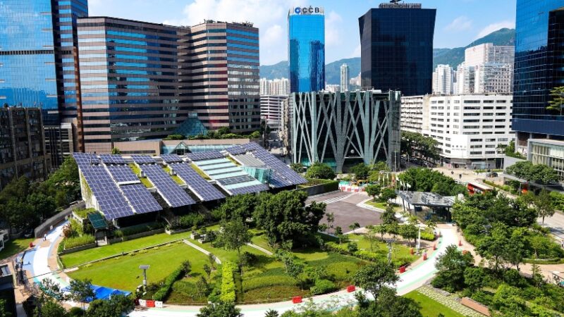 Modern sustainable building with rooftop solar panels surrounded by green park and high-rise buildings in an eco-friendly urban development.