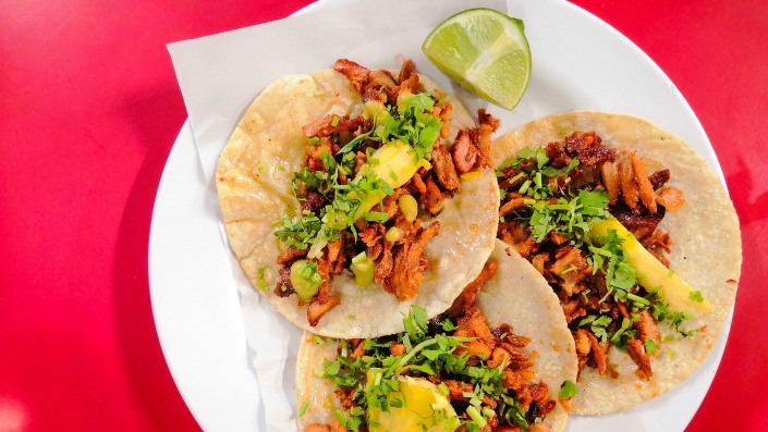 A colourful plate of Mexican tacos - corn tortillas filled with marinated meat, topped with chopped onions, cilantro and lime wedges.