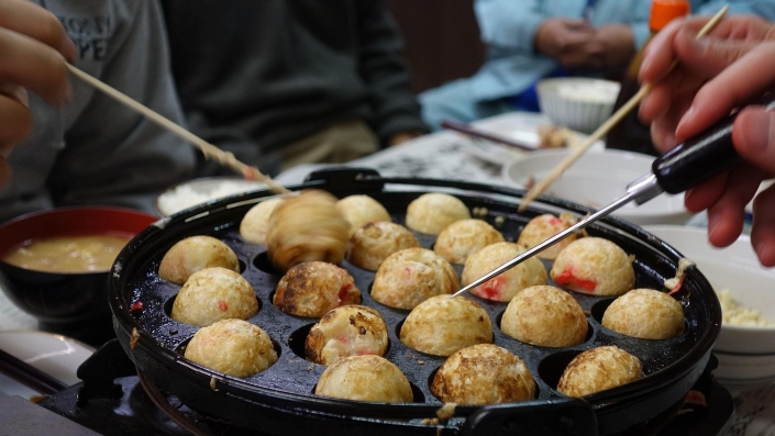 People cooking takoyaki, Japanese octopus balls, on a specialised pan with round moulds.