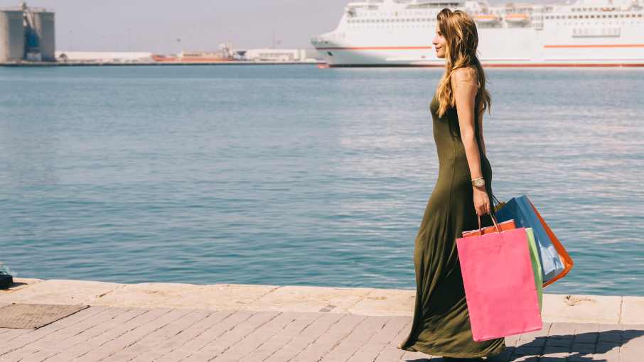 Woman walking along a marina promenade after luxury shopping, carrying designer bags in a Mediterranean setting.